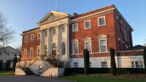 Warrington Town Hall is a large and imposing three-storey building mainly constructed from red bricks. At the centre of the building are three white columns leading up to a triangular pediment.
