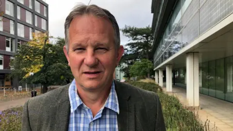 Vikki Irwin/BBC Andy Mellen is standing outside on a a raised bed of flowers between two buildings. He is wearing a Green tweed jacket with blue and white checked shirt.