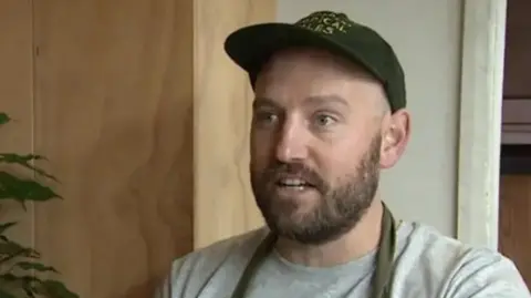 A man in a green baseball cap, wearing a pinny and grey sweatshirt looks away from the camera. He has a beard.