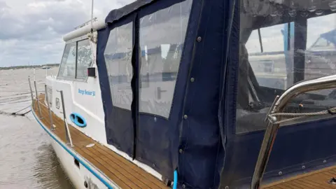 Holly Brookes A view down the side of a small houseboat that is moored on the edge of a river. The boat is painted white at the front and its rear is covered by a blue plastic sheeting with clear panels simulating windows.
