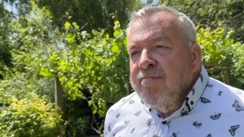 Headteacher Mark Wildman is wearing a white shirt with black butterflies and bees on it. Behind him, are a number of green trees and bushes. He has grey hair and a grey beard.