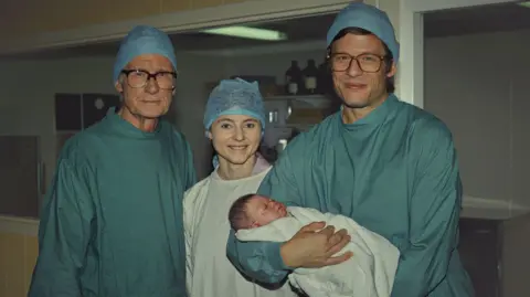 Netflix Bill Nighy, Thomasin McKenzie and James Norton are stood next to each other in blue scrubs. James is holding a baby wrapped in a white blanket.