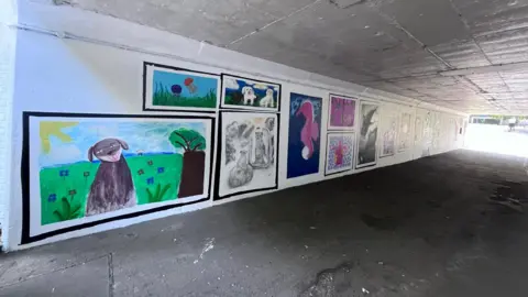 West Northamptonshire Council A long underpass, with artwork on the walls. There is a walkway in the middle. All the walls have a white base coat. 