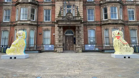 BBC Two decorated pottery dogs against the backdrop of Hanley town hall. Both are of a cream base colour. One has black fur and a red chain, the other yellow fur and a white chain.