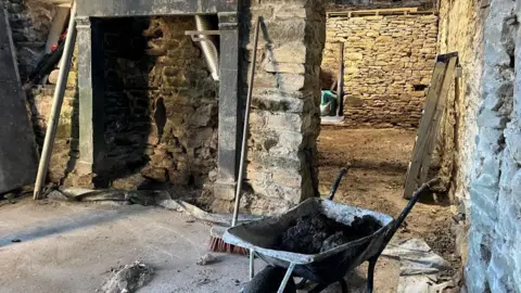 BBC/JULIA LEWIS Stone walled interior with concrete and mud floors, a mantlepiece, broom, and wheelbarrow containing mud.