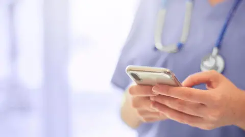 A generic picture of a doctor, who is holding a mobile phone in her hands and is wearing a stethoscope around her neck. 