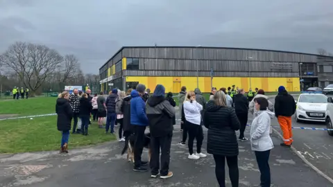 A group adults who look like they could be parents standing behind blue and while police tape across a foot path and road towards a two storey wood and yellow clad school building. About sixt police officers in high viz jackets can bee seen in a groups beside a police car about half way to the school building. Another group of four officers are together at the side entrance of the school building. It's a grey day. 
