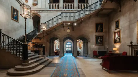 Ashridge House A grand historical stone entrance hall featuring a floating staircase, arched doorways, blue patterned rug and framed portraits mounted on the walls.