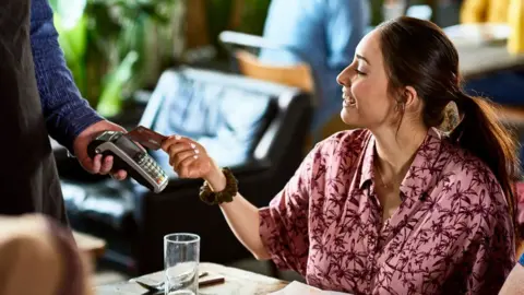 Getty Images A woman sitting in a restaurant smiles as she taps her card to pay on a card machine. 