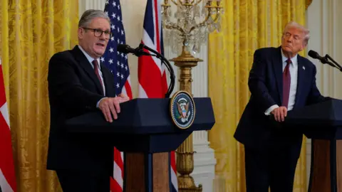 Reuters Sir Keir Starmer talks from behind a lectern during a news conference, with Donald Trump looking at him from the background. They are standing in front of a yellow curtain and a Union Jack and US flag. 