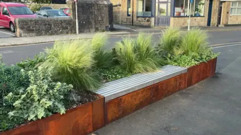 A planter with rusty walls has been completed, green plants sway in the wind and a bench for is on one side. 