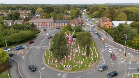 PA Media An aerial view shows a large roundabout covered in hundreds of St George's flags planted across its grassy centre. A tall silver miner statue draped in a Union Jack stands at the middle of the island with its arms raised. Cars travel around the roundabout with Brownhills Community Centre and residential streets visible beyond.

