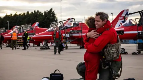 A member of the red arrows hugging a child, the man is smiling with his eyes closed.