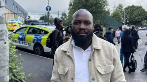 A man in a white polo shirt and beige jacket stands on a pavement. There are police cars in the background.