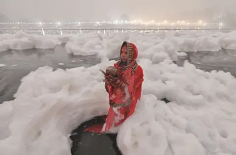 Somenath Mukhopadhyay A devotee stands in the Yamuna River, New Delhi, offering prayers to the sun, as the water - thick with foam - surrounds her. She is wearing a red sari