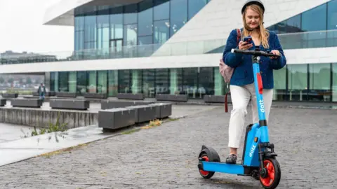 Dott A woman looking at her phone with one foot on a blue e-scooter, which has the name "dott" printed on.