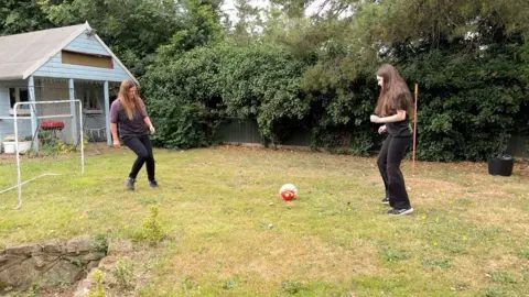 Two men women kick a ball around a garden. A shed and goalposts can be seen in the background, alongside trees.