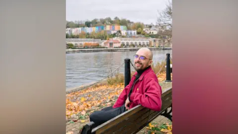 University of Bristol Jacob is sitting on a bench beside Bristol Docks with a multi-coloured row of terraced houses on the hill on the further bank. He is looking towards the camera over his left shoulder and is wearing a bright pink jacket and smiling