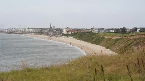 A curving sandy beach sitting below grassy cliffs. Small figures walk along the shoreline beside grey-ish water. A coastal town with low buildings and a tall church spire rising above the rooftops sits behind the beach. The sky is overcast. 