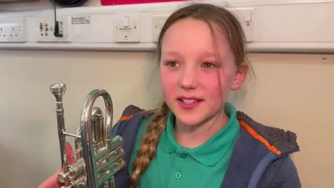 May Birkett has with her dark blonde hair in a plait and is wearing a green polo shirt and blue hoodie. She is holding a trumpet and is standing against a wall. 