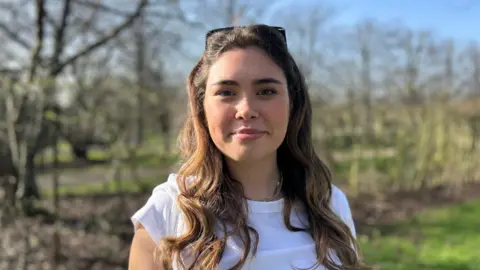 Woman with long brown hair, white t-shirt, looking straight at the camera, she has her hair pulled back with a pair of sunnies and in the background there's a line of trees and grass.