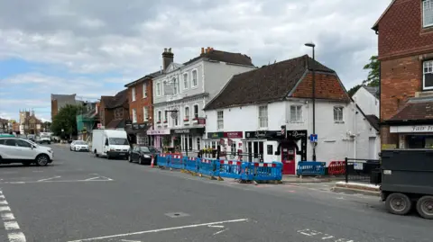 BBC Marlborough High Street, with shops and vehicles along the main road. Blue work barriers are in front of three shops. 