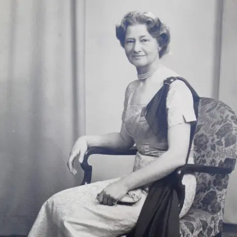 Stephen Artus A black and white photo of a lady sat in an arm chair with a long dress on