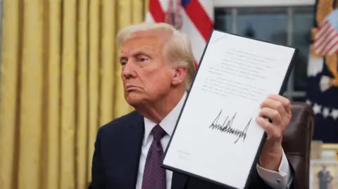 President Donald Trump holds a document he signed while he sits in the Oval Office. His expression is neutral and he is in a dark suit and tie. 