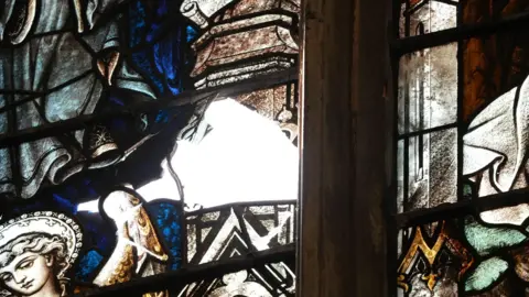 BBC News/Jasmine Lowe This image is a close up shot of the hole in the stained glass. An angel is depicted next to the hole. 