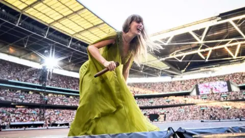 Getty Images Singer Taylor Swift in a long green dress holding a microphone crouching down smiling while on stage with the crowd behind her at Wembley Stadium in London