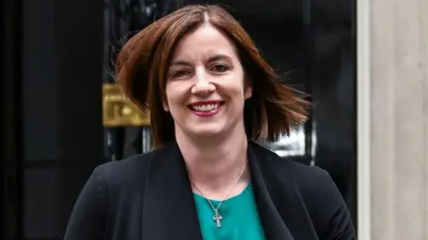 Phillipson wears a teal scoop-necked top with a black jacket and a small silver Celtic cross. She wears dark pink lipstick and is smiling. Her light brown hair is worn loose, straight and shoulder-length.