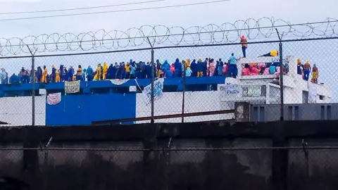 Inmates dressed in prison jumpsuits can be seen during a protest at the prison in Barinas state. They have hung sheets bearing messages from the roof.