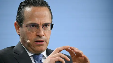 Getty Images A man with black gelled back hair speaks into a microphone attached to his face. He is wearing glasses and a dark suit, white shirt and blue tie.