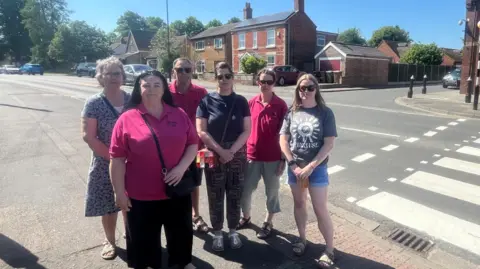 Six people stand in a group by a zebra crossing. One woman in the back in a blue and white floral dress; a woman in a dark pink T-shirt and black trousers; one man in a dark pink T-shirt; a women in a navy T-shirt and printed leggings; another woman in a dark pink T-shirt and green trousers; a women in a grey T-shirt and blue denim shorts. 
