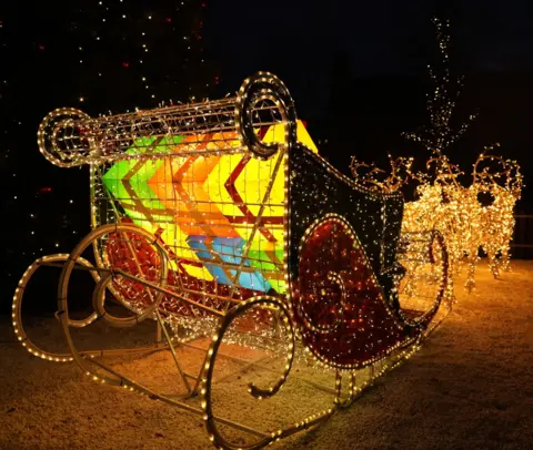 Rosie McGeachan Large sleigh ornament illuminated with fairy lights with brightly coloured and lit gifts inside, led by LED lit reindeers.