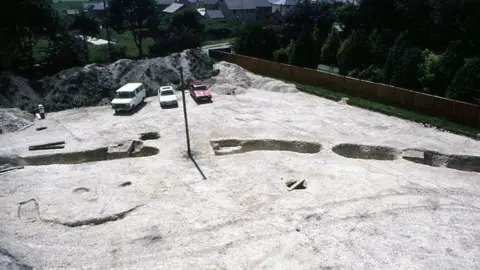 Dorset Museum Aerial view of the original excavation. Three cars are parked on the side. It's a sunny day.