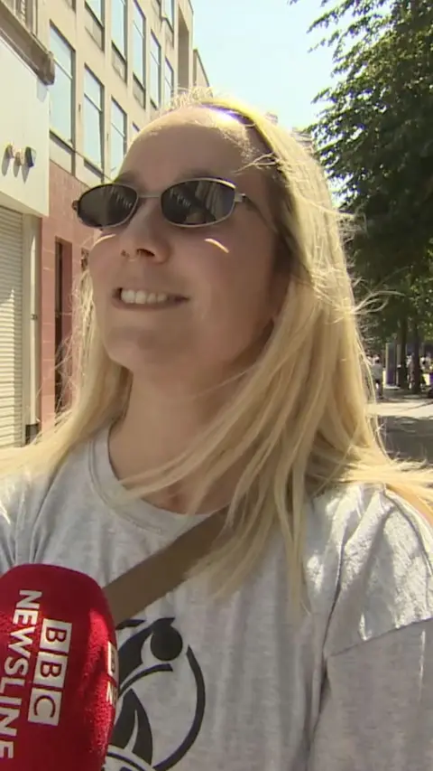 A woman with long blonde hair wearing black sunglasses and a grey tshirt. She is smiling with a red microphone with BBC Newsline written in white on it. 