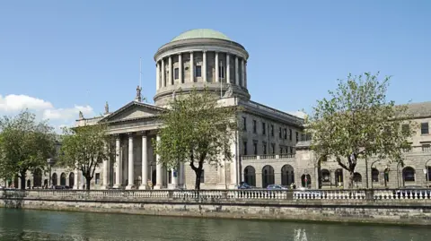 Getty Images A wide shot of Dublin's High Court, showing a large white stone building with a large dome top. In front of the building is the River Liffey with four trees set up along the river's edge. The sky is blue and there are a few small clouds on the left hand side of the image.