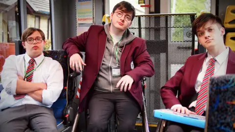 BBC / Tiger Aspect & Testmouse Prod / Sam Ward 'Dan' (Reuben Reuter), 'Mike' (Jack Carroll), 'Sonny' (Zak Ford-Williams) on a small bus, they're all in school uniforms, and looking at the camera passively