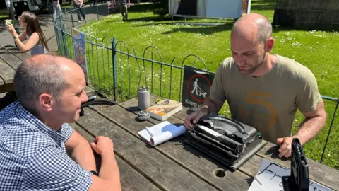 Patrick and Arek sit at a park bench together working on a poem. Patrick is rolling a piece of paper onto his typewriter and Arek is looking at him.