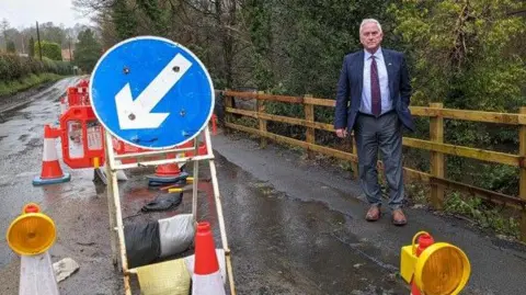 Glen Sanderson at the landslip on the B6343 at Mitford near Morpeth.
He is standing next to wooden fence on the right and wears a suit and a tie.
Water is running down the centre of the road with traffic cones surrounding the small stream. A blue circle sign with a white arrow pointing to the left is put in the middle of the road.