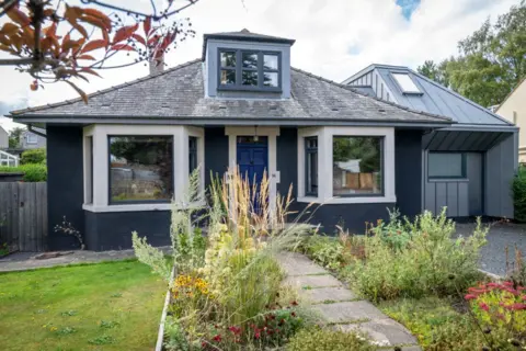 IWC Media Homegrown Hoose. A dark coloured bungalow with two bay windows at the front and a small garden.