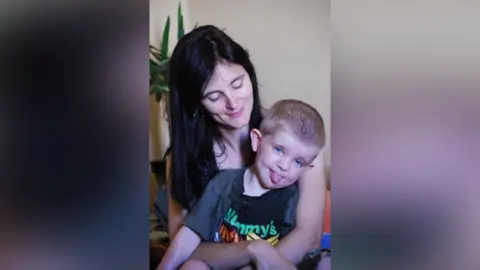 PA Media Elizabeth Moate has long dark hair and she is looking down lovingly at her son, who she is holding on her lap. He has fair cropped hair and is sticking his tongue out. His is wearing a black T-shirt with a graphic that partly says 'Mummy's'.