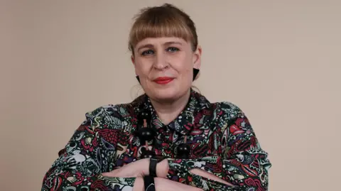 A woman with blonde hair tied back. She is wearing a multi-coloured patterned top and has her arms folded. 