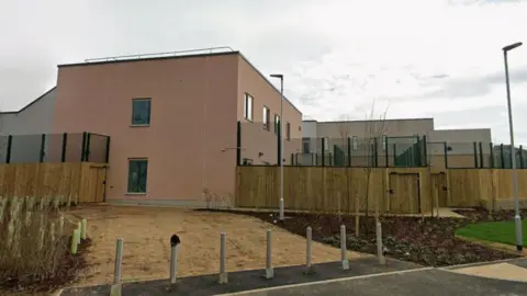 The image shows the outside of a mental health trust: wooden beams and green gates can be seen on the exterior and the outside of the building is painted a creamy pink.