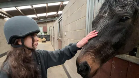 In a stable, built from breeze blocks, a chestnut coloured horse is being petted on the head by a girl wearing a grey riding helmet.