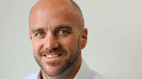 A bearded balding man with a blue checked shirt.