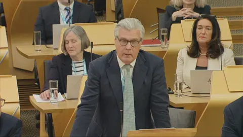 Scottish Parliament Malcolm Offord, wearing a dark suit and green tie, stands to speak in the Scottish Parliament