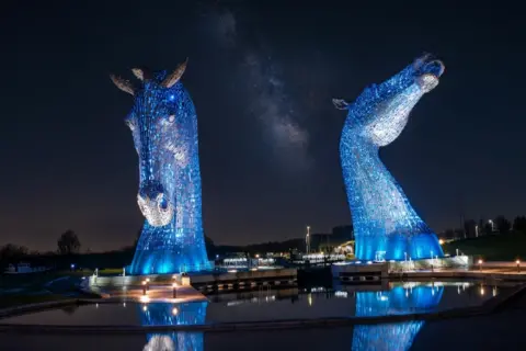 Gordon Stewart The Kelpies statues lit up blue at night. The sky is dark and there are street lights at the bottom of the statues.