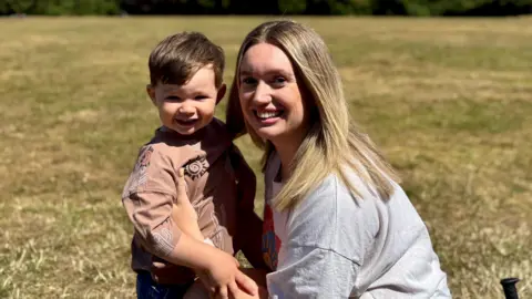 Karis in a grey T-shirt, holding two-year-old Noah, standing on a lawn, both are smiling